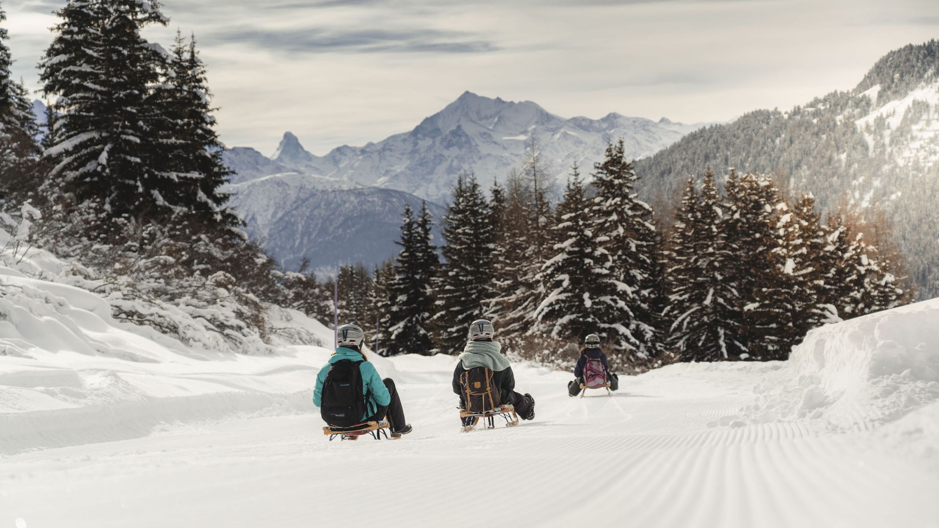 Auf der Moosfluh bei Riederalp fahren drei Personen mit dem Schlitten auf einer frisch präparierten Piste durch den verschneiten Bergwald.