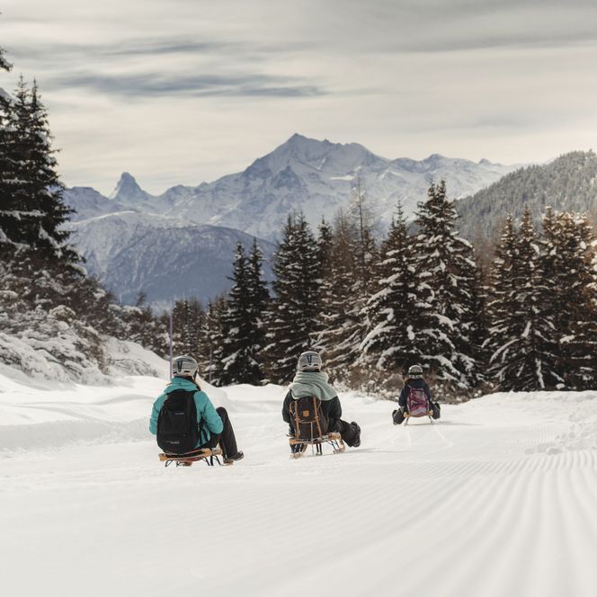 Auf der Moosfluh bei Riederalp fahren drei Personen mit dem Schlitten auf einer frisch präparierten Piste durch den verschneiten Bergwald.