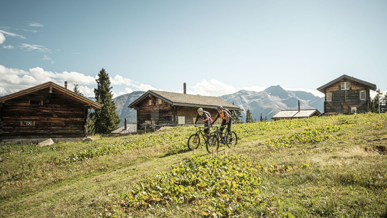 Zwei Mountainbiker fahren bei der Fiescheralp an Chalets vorbei durch alpine Wiesen in der Aletsch Arena
