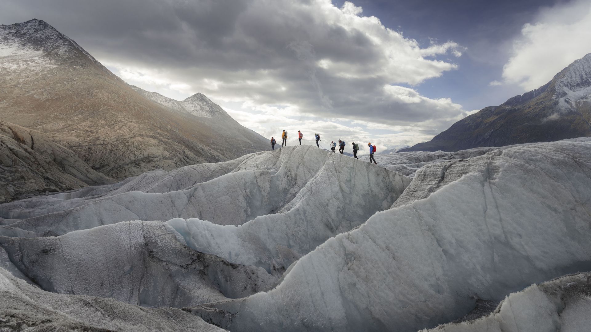 A group of hikers walk one behind the other over a white ice ridge. This is how close you can experience the Great Aletsch Glacier on tours.