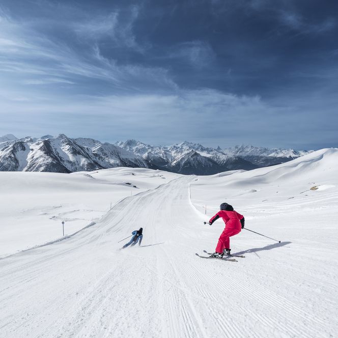 Deux personnes sur une piste extrêmement large et ensoleillée devant un grand panorama de montagnes en faisant du ski dans l'Aletsch Arena.
