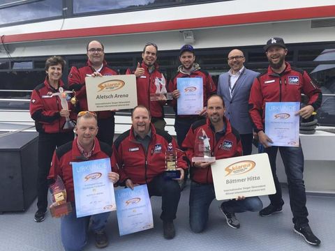 Auszeichnungen Aletsch Arena: Gruppe mit Skiareatest-Urkunden und Trophäen vor einer Bergbahn.