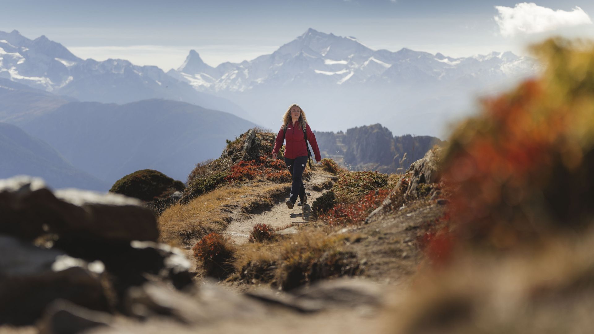 A woman in a red jacket on a ridge path, a colorful and sunny experience while hiking in the Aletsch Arena in autumn.