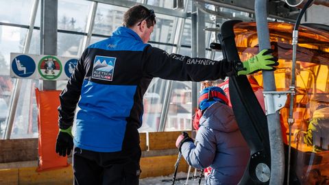 Jobs Aletsch Bahnen AG: Bahnpersonal unterstützt eine Person mit Helm und Skistöcken beim Einstieg in die Gondel.