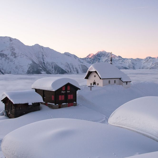 La chapelle enneigée Maria zum Schnee se dresse avec des chalets dans l'hiver Aletsch Arena au-dessus d'une épaisse mer de nuages.