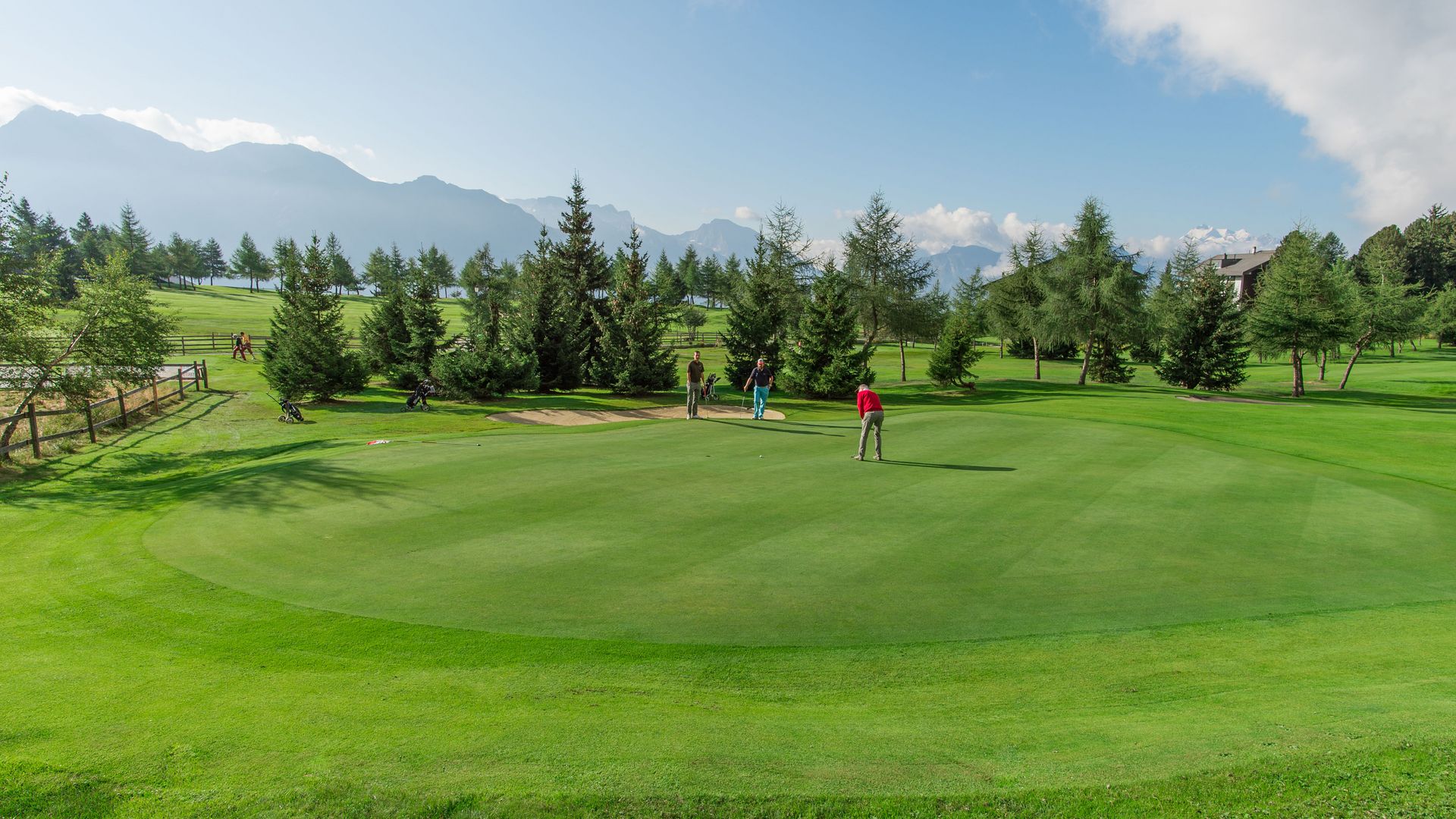 Plusieurs golfeurs jouent sur le green impeccablement entretenu du Riederalp Golf, entouré d'arbres et d'un panorama montagneux impressionnant.