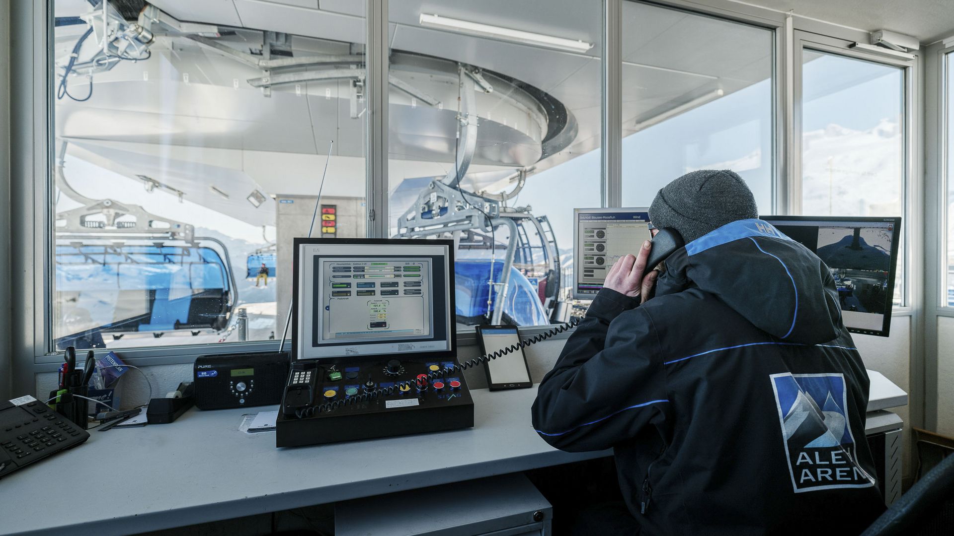 Jobs Aletsch Bahnen AG: Person telefoniert am Steuerpult vor Monitoren mit Blick auf Gondeln in der Station.