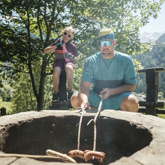A father grills sausages over a fire in Lax Valais, while his daughter, dressed in cycling gear, sits laughing in the background.