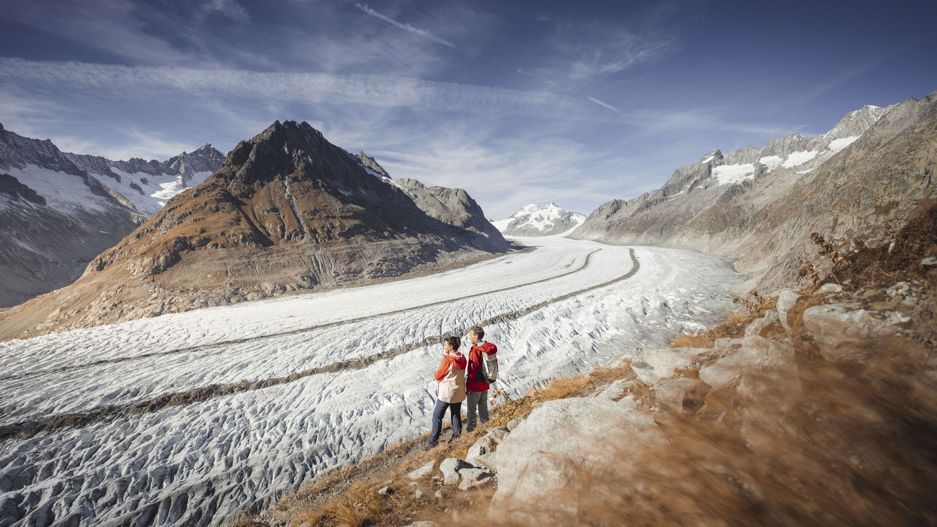 A pair of hikers enjoy the direct view of the Grosser Aletsch Glacier in the warm light from the rocky vantage point.