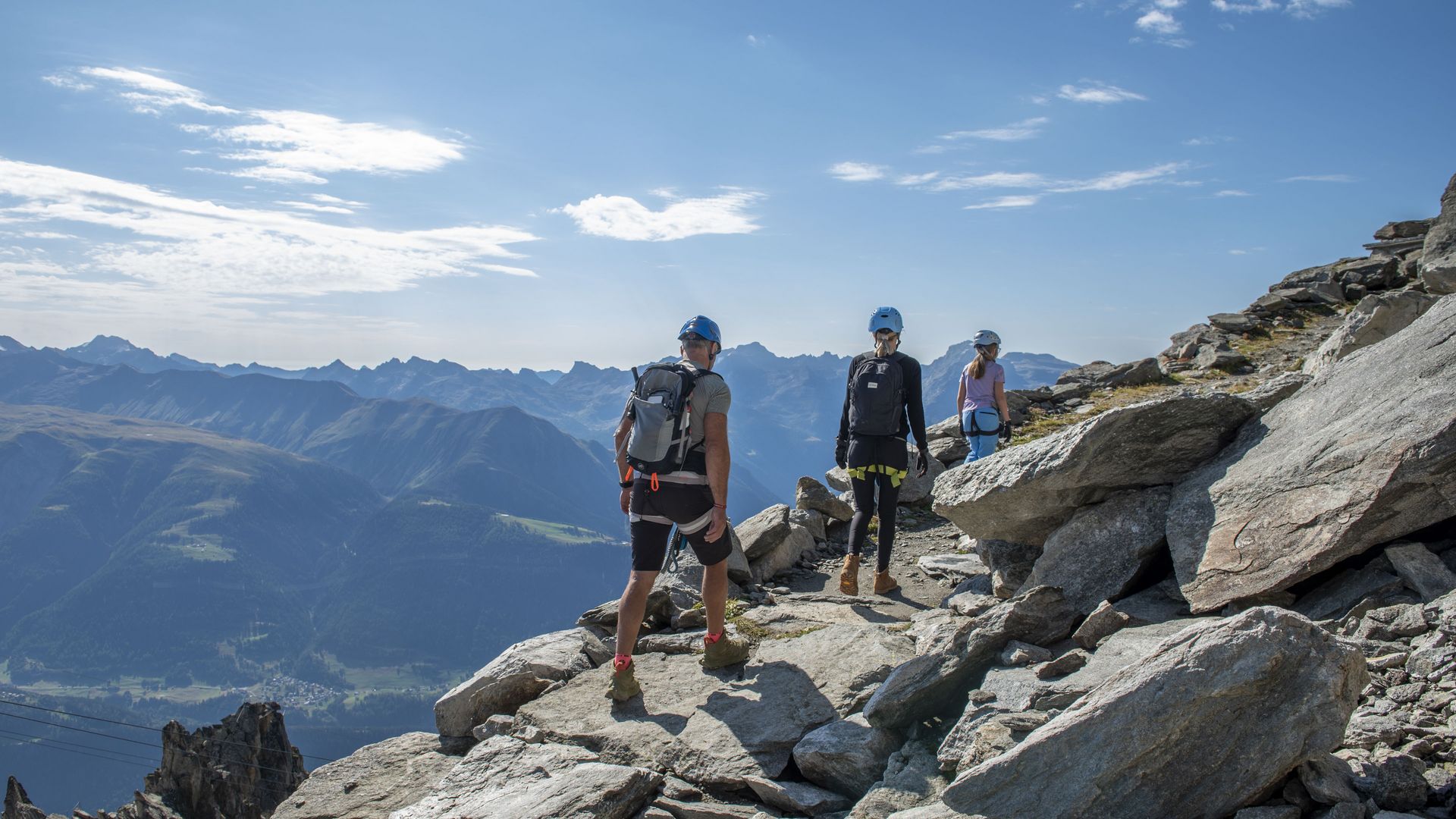 Une famille équipée de casques randonne sur un terrain rocheux le long d'une via ferrata et découvre l'escalade dans l'Aletsch Arena, au-dessus des Alpes.