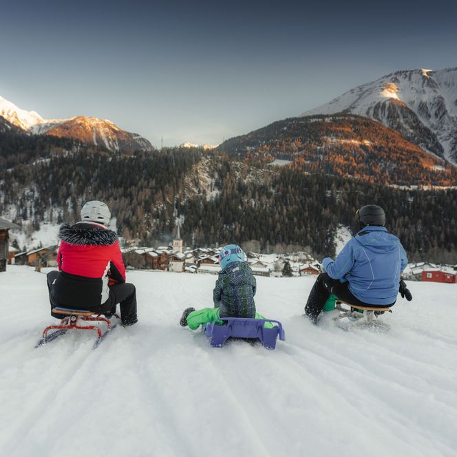 A family of three sits on sledges and looks down on the snow-covered village of Lax Valais in the evening sun.