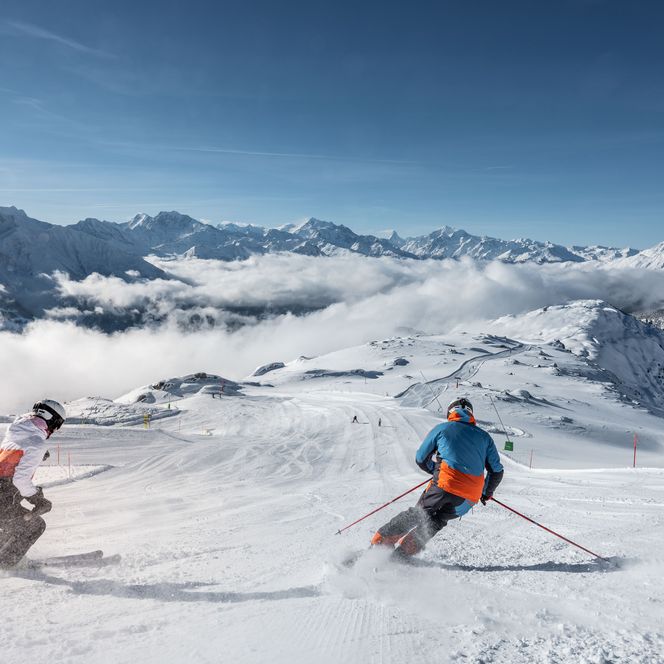 Deux skieurs dévalent une piste par un temps radieux, directement en direction d'une épaisse mer de nuages dans l'Aletsch Arena d'hiver.