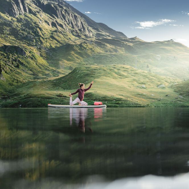 Eine Frau praktiziert SUP Yoga auf dem Bettmersee in der Aletsch Arena, umgeben von Bergen und ruhigem Wasser.