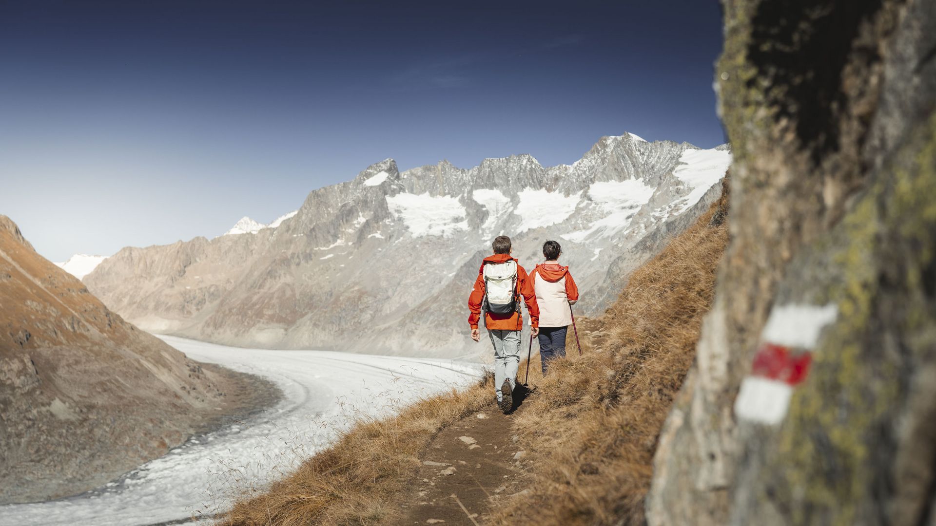 A couple hiking in the Aletsch Arena on a rocky mountain path, right next to the mighty Aletsch Glacier.