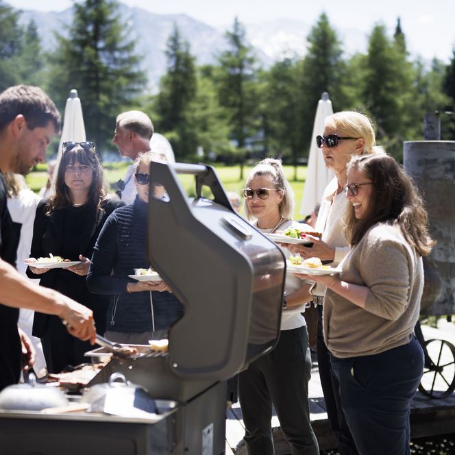 Lachende Menschen holen sich während dem Retreat Aletsch Arena draussen mit Tellern in der Hand Essen vom Grill.