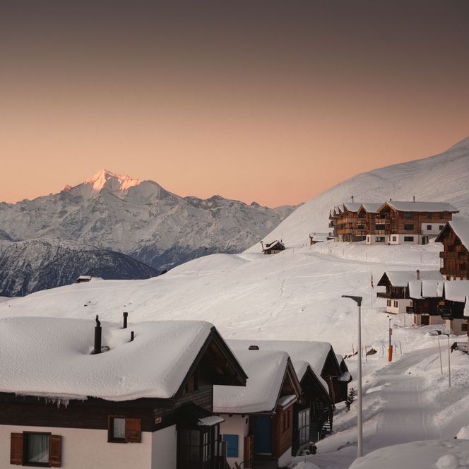 Chalets enneigés le matin, tandis qu'en arrière-plan, un sommet de montagne s'illumine de rouge dans l'Aletsch Arena d'hiver.