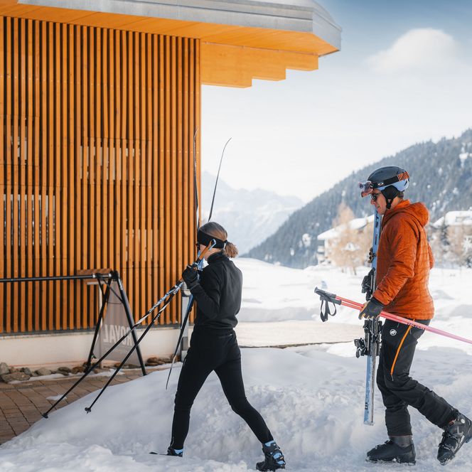 Ein Mann und eine Frau tragen ihre Skier bei einem Retreat Aletsch Arena im Winter an einem Gebäude vorbei.