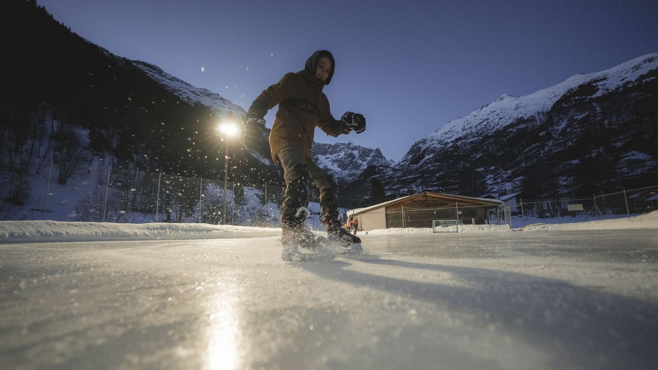 Ein Kind läuft auf der Natureisbahn „Inschi Ischbahn” im Fieschertal Schlittschuh, während im Hintergrund die verschneiten Berge in der Abenddämmerung zu sehen sind.
