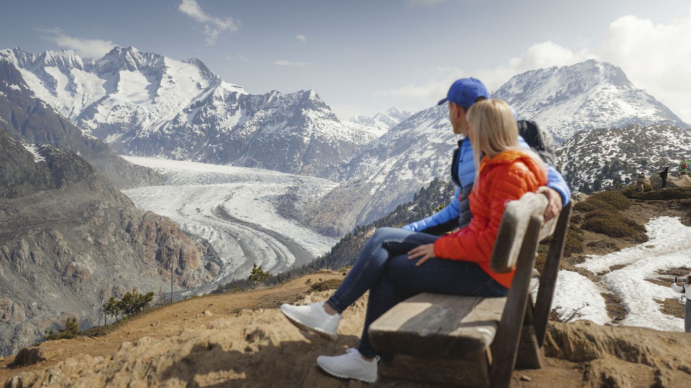 Couple assis sur un banc en bois, regardant l'impressionnant glacier d'Aletsch et les montagnes enneigées.