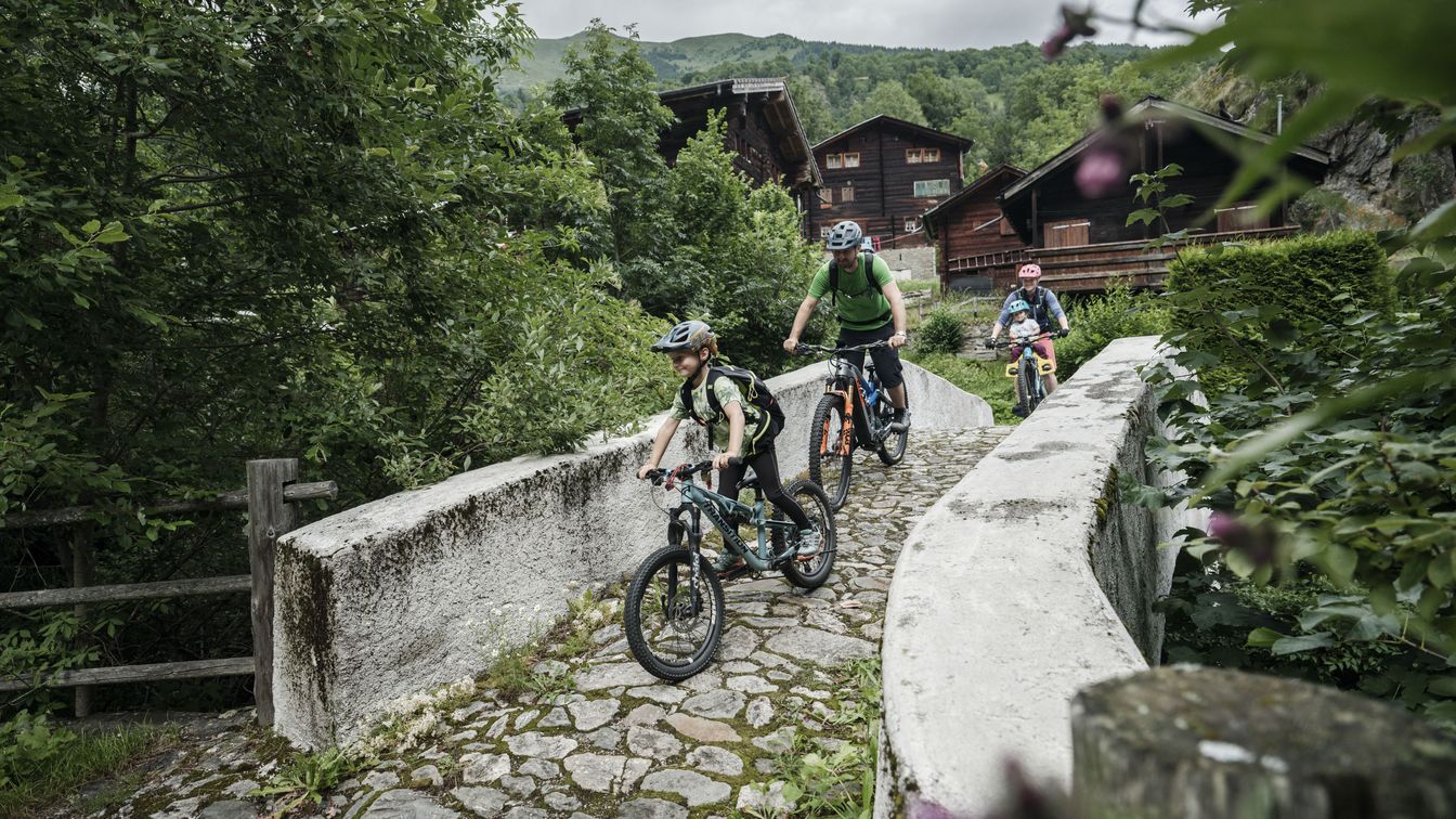 Eine Familie fährt mit Mountainbikes über eine Steinbrücke, die in der Nähe von traditionellen Walliser Holzhäusern im Fieschertal liegt.