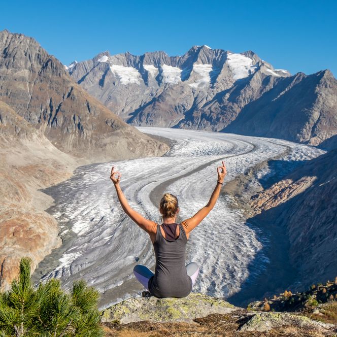 Eine Frau meditiert beim Yoga mit erhobenen Armen über dem Grossen Aletschgletscher in der Aletsch Arena.