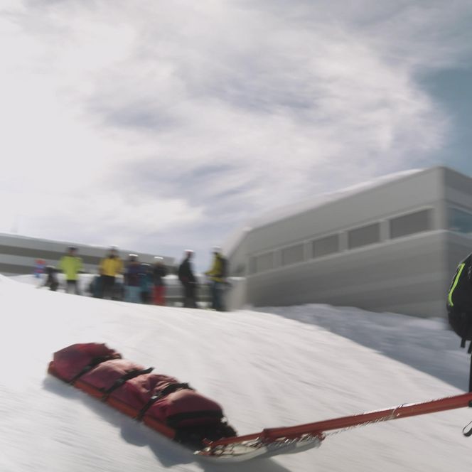 Jobs Aletsch Bahnen AG: Pistenrettung zieht einen roten Rettungsschlitten auf Skiern an einer Bahnstation vorbei.