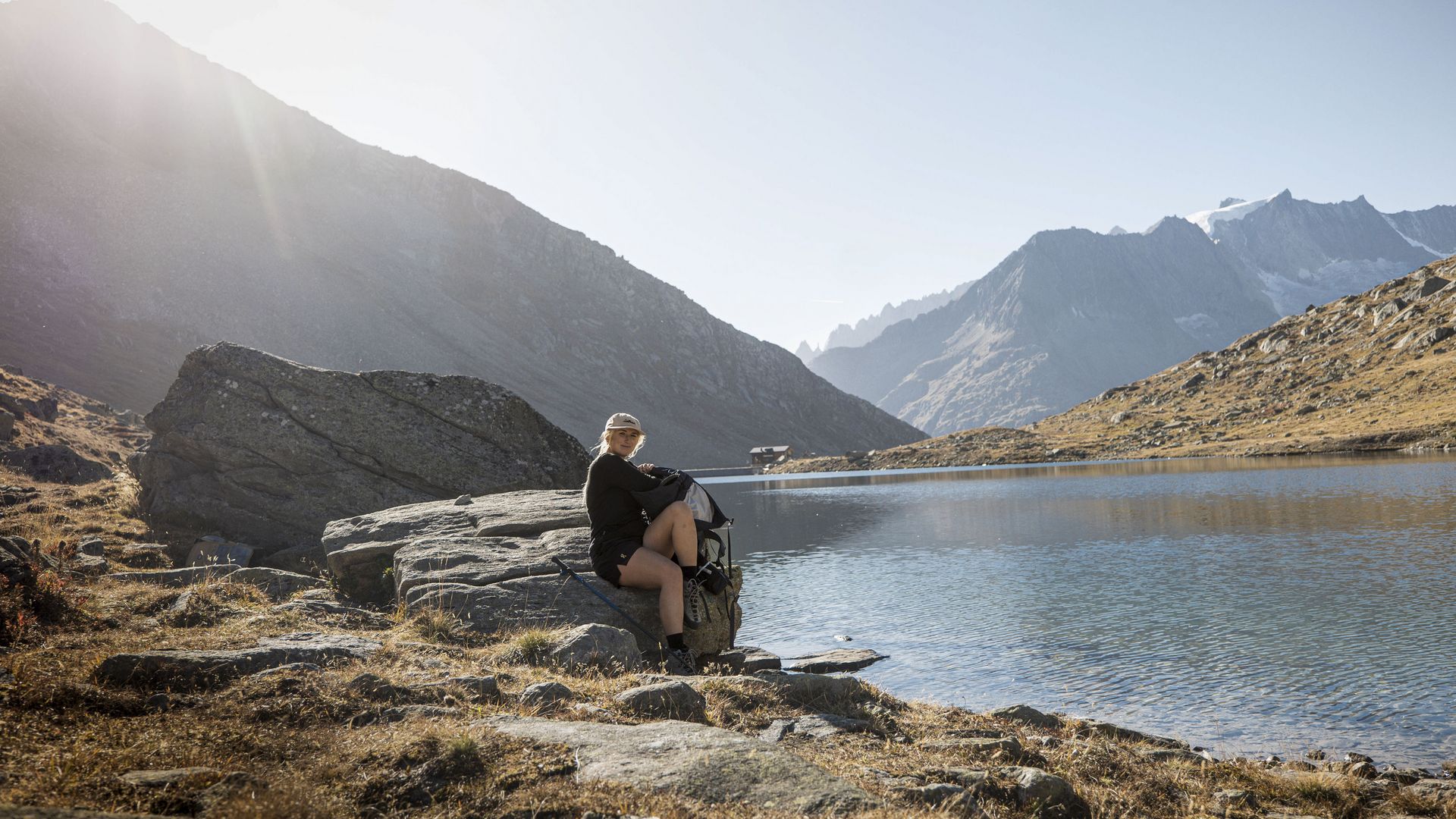 A young woman with a rucksack sits by a blue mountain lake, an ideal break while hiking in the Aletsch Arena.