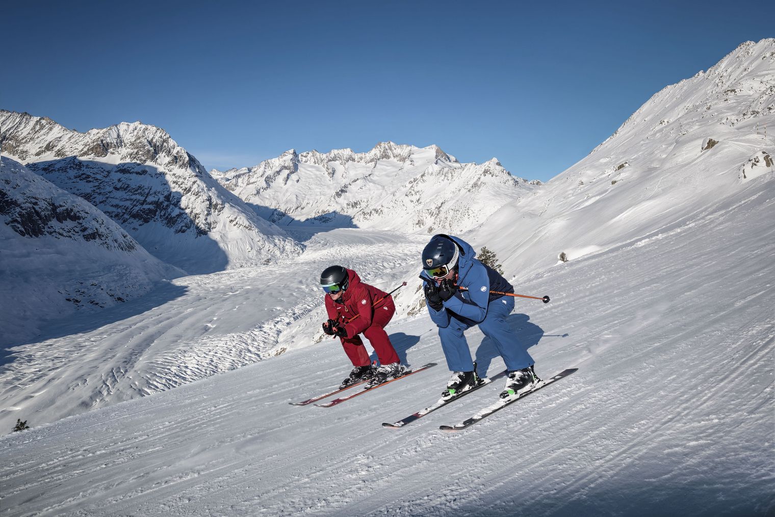 Deux skieurs dévalent la piste à toute vitesse, profondément accroupis, dans l'hiver de l'Aletsch Arena, avec en arrière-plan l'immense coulée de glace qui se repose.