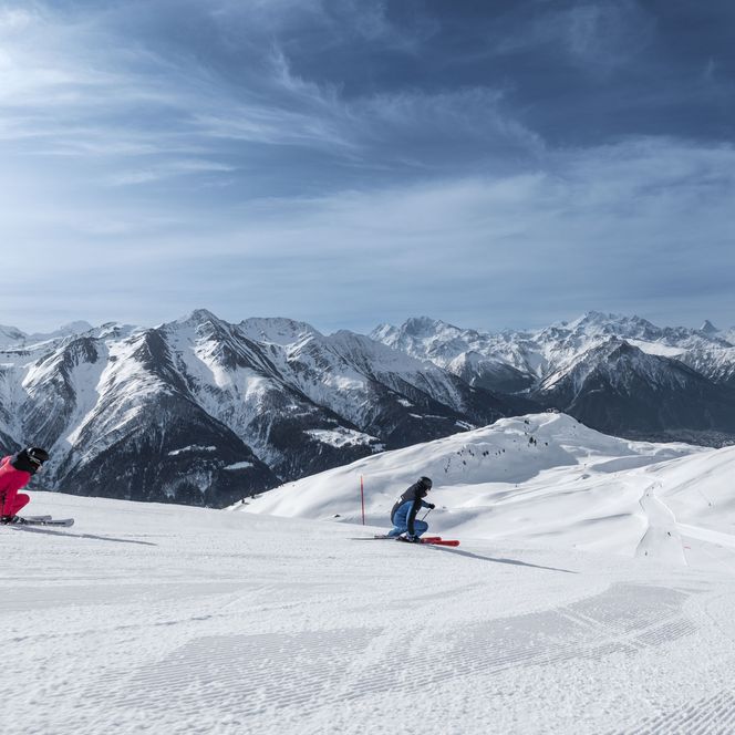 Deux personnes sportives skient sur une piste de ski extrêmement large et ensoleillée devant un grand panorama de montagnes dans l'Aletsch Arena d'hiver.