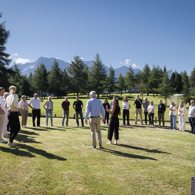 Eine Gruppe steht während dem Retreat Aletsch Arena im Kreis auf einer sonnigen Wiese vor einer Berglandschaft.