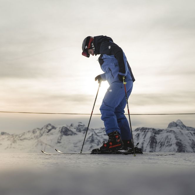 Un skieur en combinaison bleue regarde ses skis dans un doux contre-jour, prêt à dévaler les pentes de l'Aletsch Arena d'hiver.