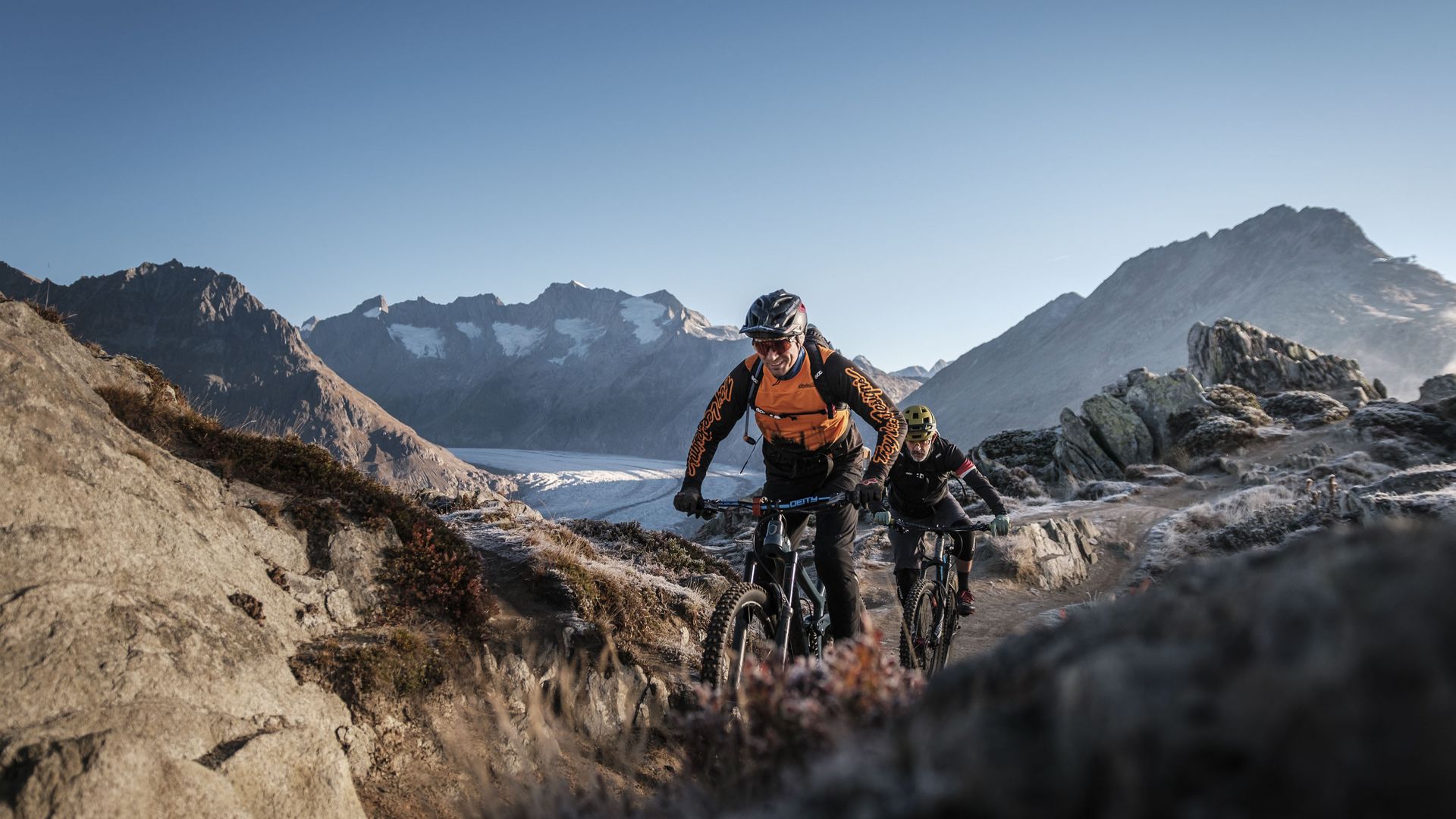 Deux vététistes parcourent en été un sentier de montagne caillouteux au-dessus de Riederalp Suisse, avec le glacier derrière eux.