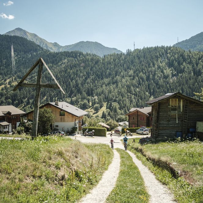A family rides mountain bikes along a sunny country lane past a large wooden cross in Lax, Valais.