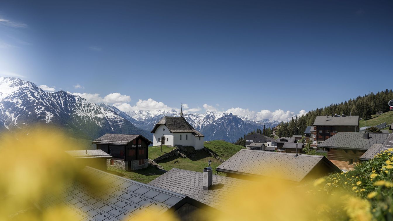 Chapelle Maria zum Schnee, entourée de fleurs, de chalets et de montagnes sous un ciel bleu.