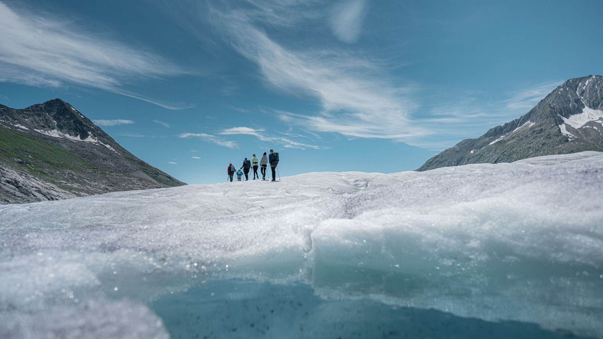 A group of mountains hiking in the distance. In the foreground, the Great Aletsch Glacier reveals deep blue ice and small pools of water.