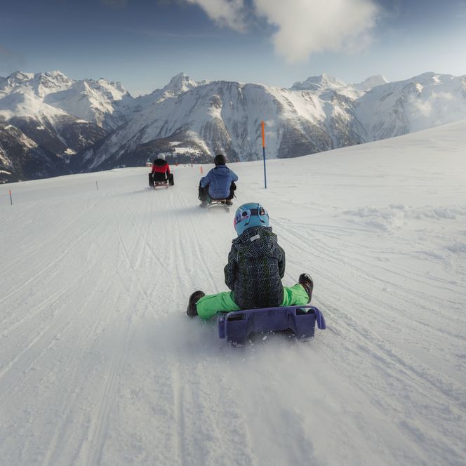 A child rides a purple sledge down a wide snowy slope in the Lax Valais region, with snow-covered mountains in the background.