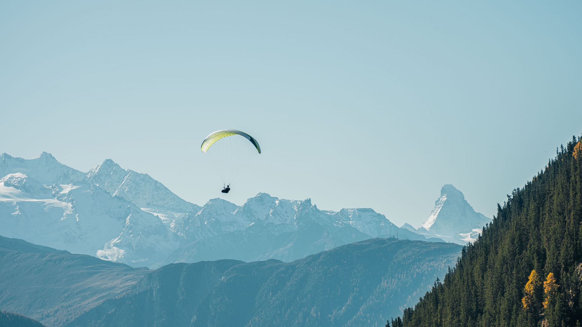 Parapente devant un imposant décor alpin et une pente boisée sous un ciel dégagé