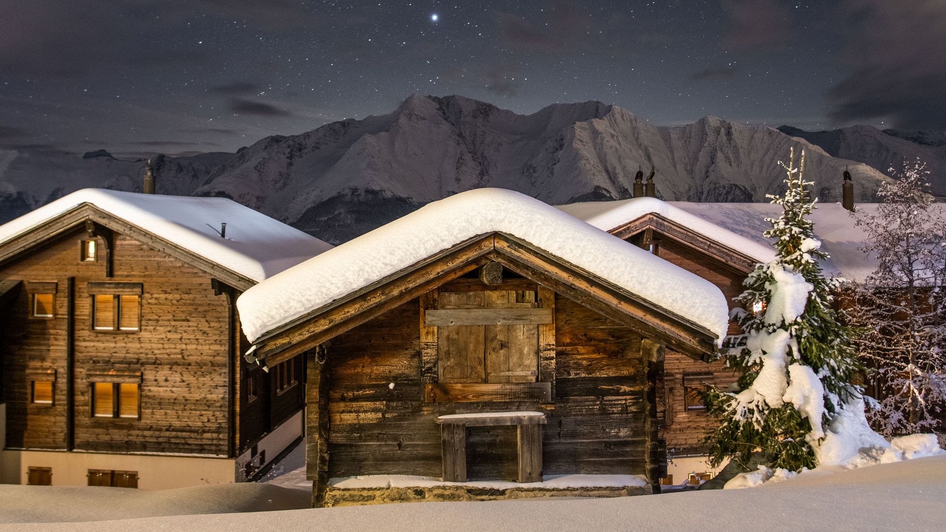 Des chalets en bois traditionnels profondément enneigés sur la Riederalp Suisse s'illuminent la nuit sous un ciel étoilé clair.