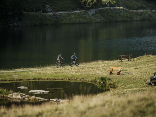 Zwei Personen fahren als Ausgleich zum Coworking Riederalp mit Mountainbikes an einem ruhigen Bergsee entlang.