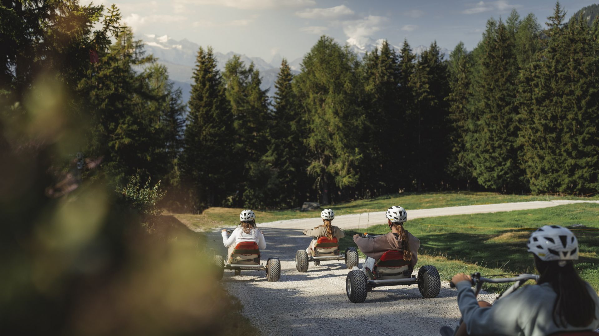Un groupe sur le mountaincart Aletsch Arena roule sur un chemin de gravier clair en direction d'une forêt de sapins.