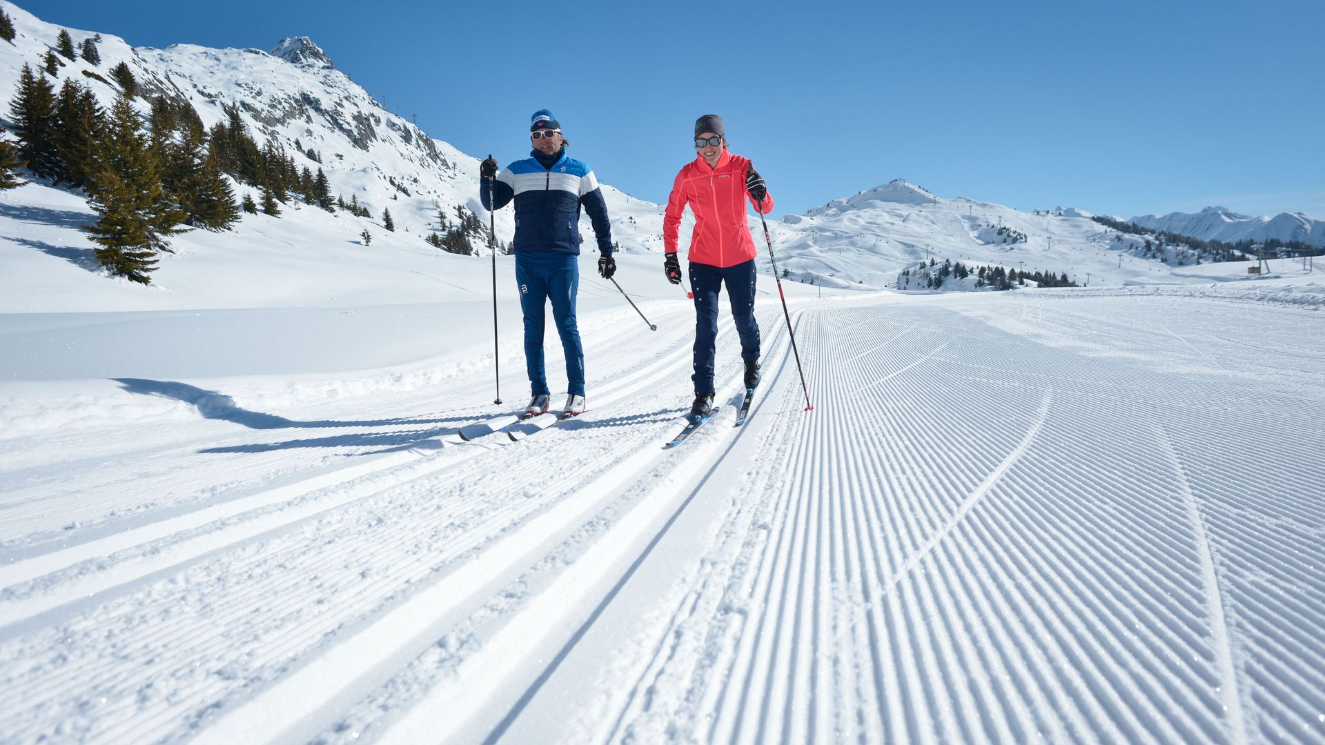 On voit deux personnes pratiquant le ski de fond en technique classique sur une piste fraîchement damée dans l'Aletsch Arena.