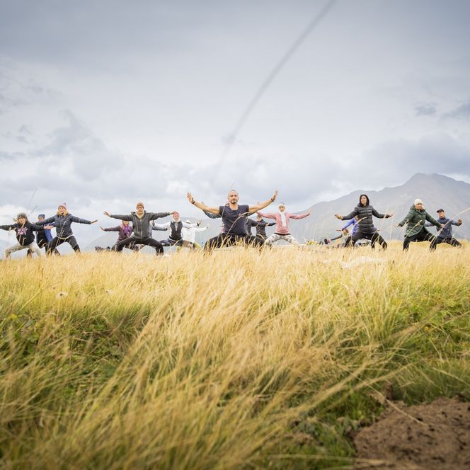 Teilnehmende des Mountain Glow Yoga Festivals üben gemeinsam auf einer Bergwiese in der Aletsch Arena vor alpiner Kulisse.